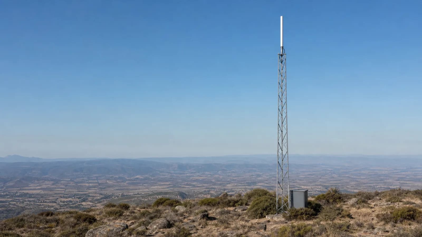 A white vertical repeater antenna on a steel lattice tower on a ridge top, with a hazy valley stretching to distant hills below