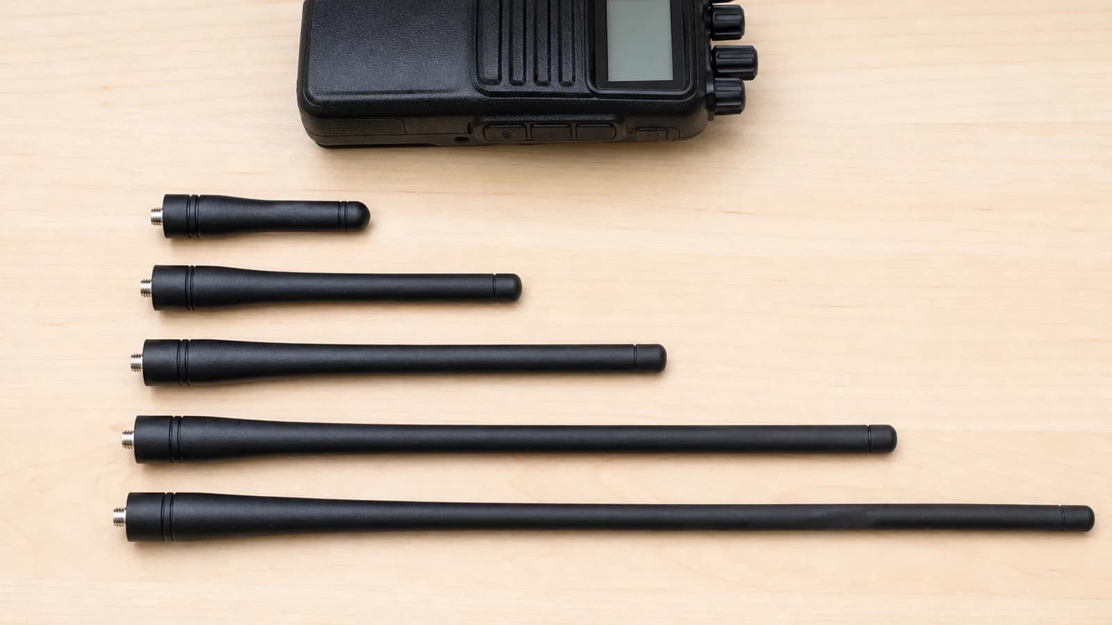 Several black handheld radio antennas of different lengths laid out side by side next to a handheld radio