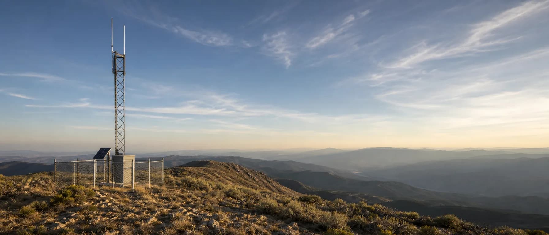 A solar-powered UHF repeater antenna mast on an open ridgeline in late-afternoon light, layered ridges fading into haze below